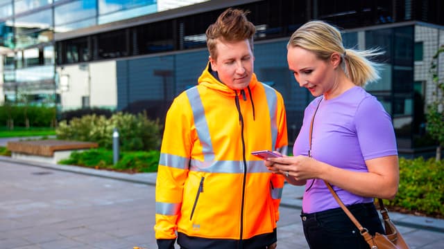 Nainen seisoo kerrostalon etuoven edessä huoltohenkilön kanssa ja tutstuu digitaaliseen huoltopalveluun