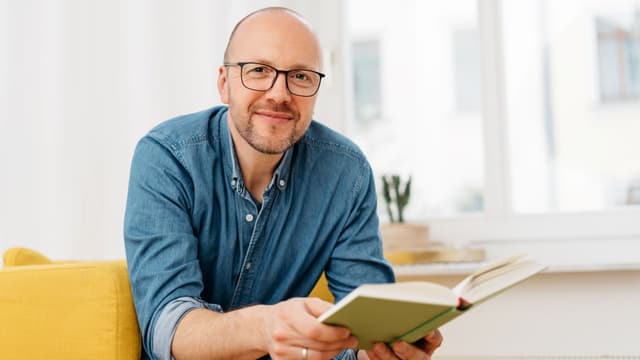 Silmälasipäinen mies istuu ja pitää avointa opaskirjaa kädessään katsoessaan suoraan kameraan.