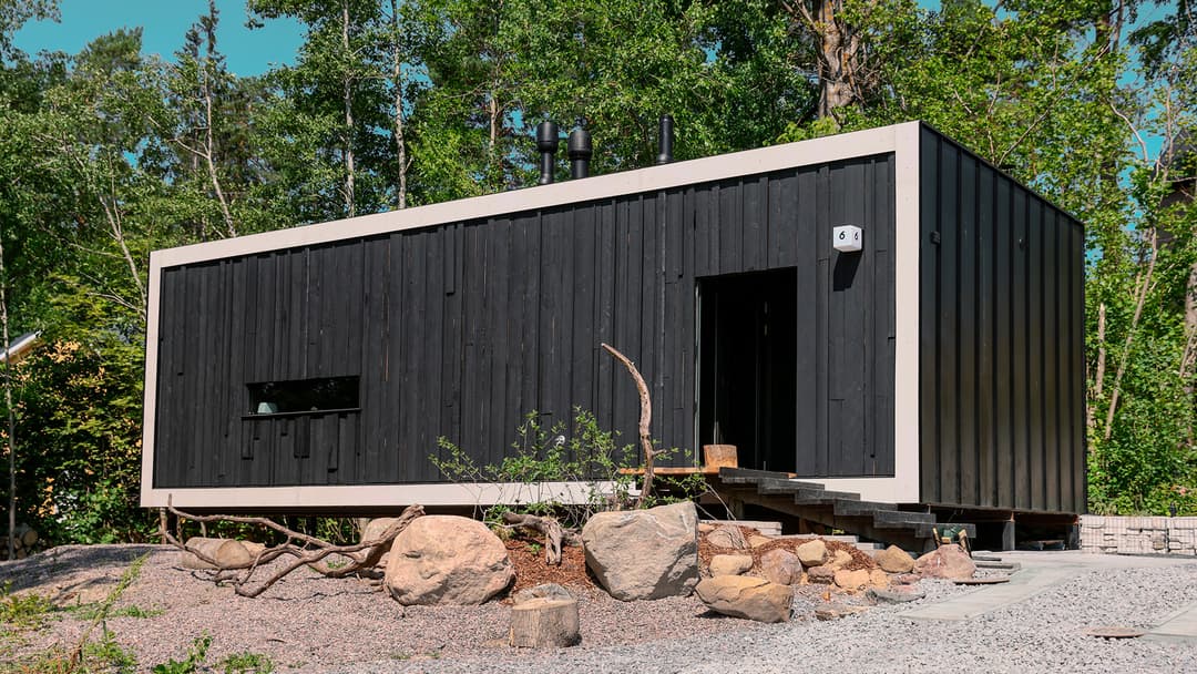 minitalo talo varjon julkisivukuva. Mustalla laudalla vuorattu merikontin kokoinen ja muotoinen minitalo. Julkisivussa on yksi pitkä ja matala ikkuna